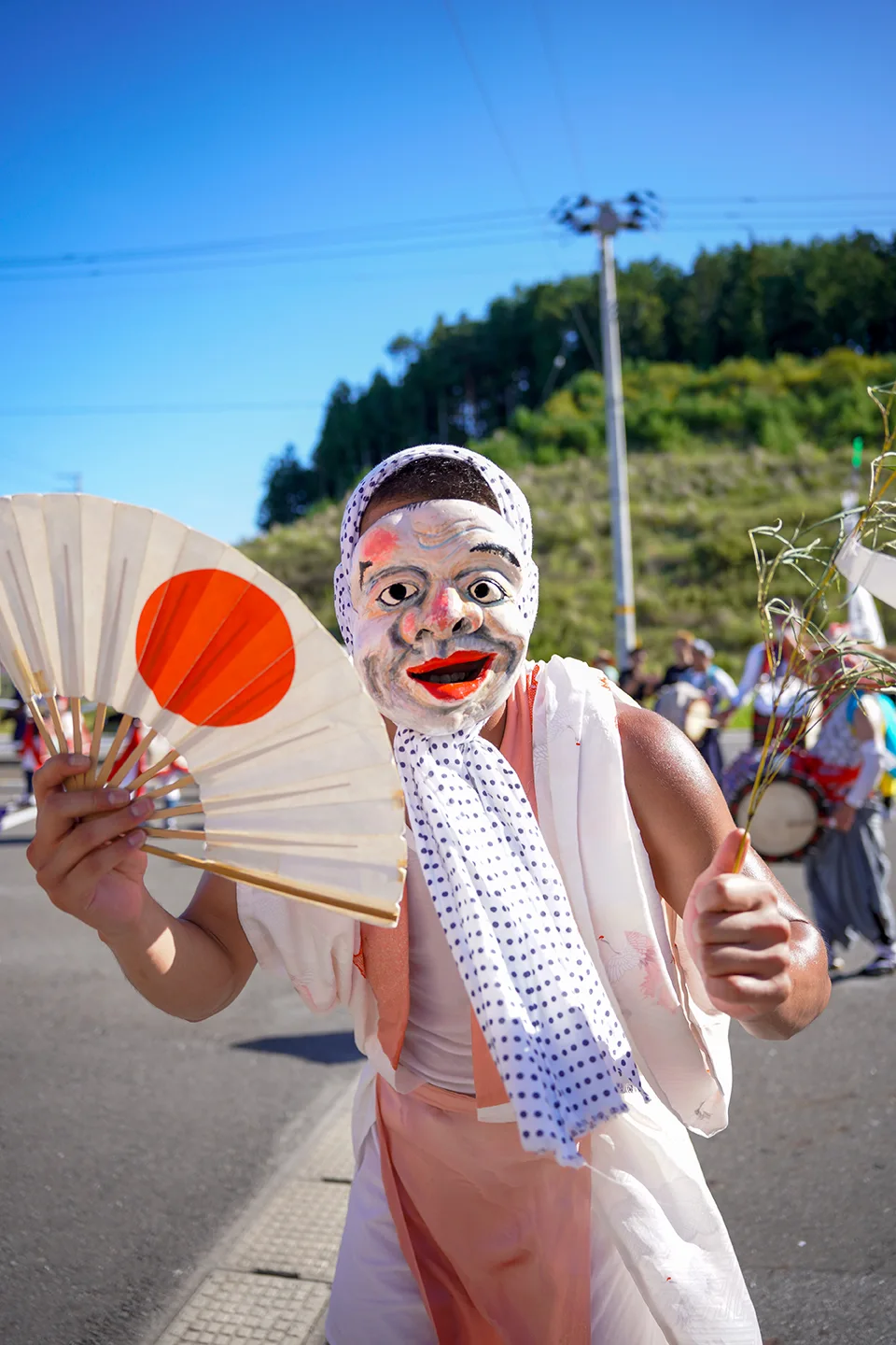 山田祭りで日の丸の扇を手に踊るお面姿の道化役